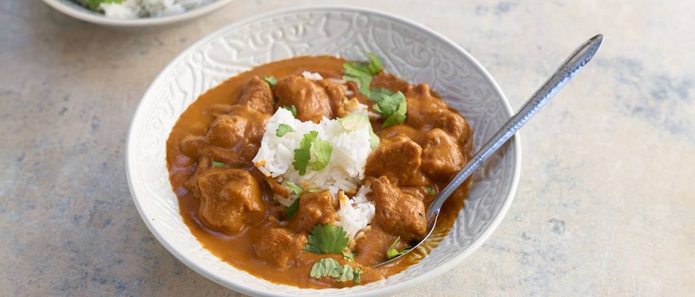 Das Bild zeigt eine Schüssel mit vegetarischem Butter-Chicken mit einer cremigen, braunfarbenen Sauce. Das Gericht ist mit weißem Reis garniert und mit frischen Korianderblättern dekoriert. Ein silberner Löffel liegt in der Schüssel. Im Hintergrund ist eine weitere Schüssel mit Reis zu sehen.