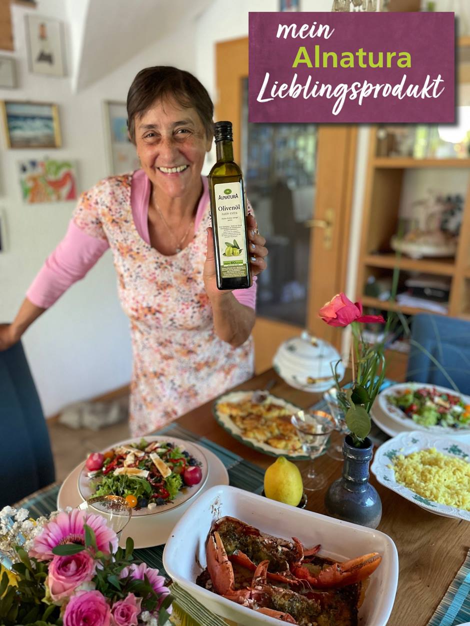Alnatura Kundin Marcia steht lächelnd vor einem Tisch voller unterschiedlicher Leckereien. In die Kamera hält sie ihr Alnatura Lieblingsprodukt Olivenöl.