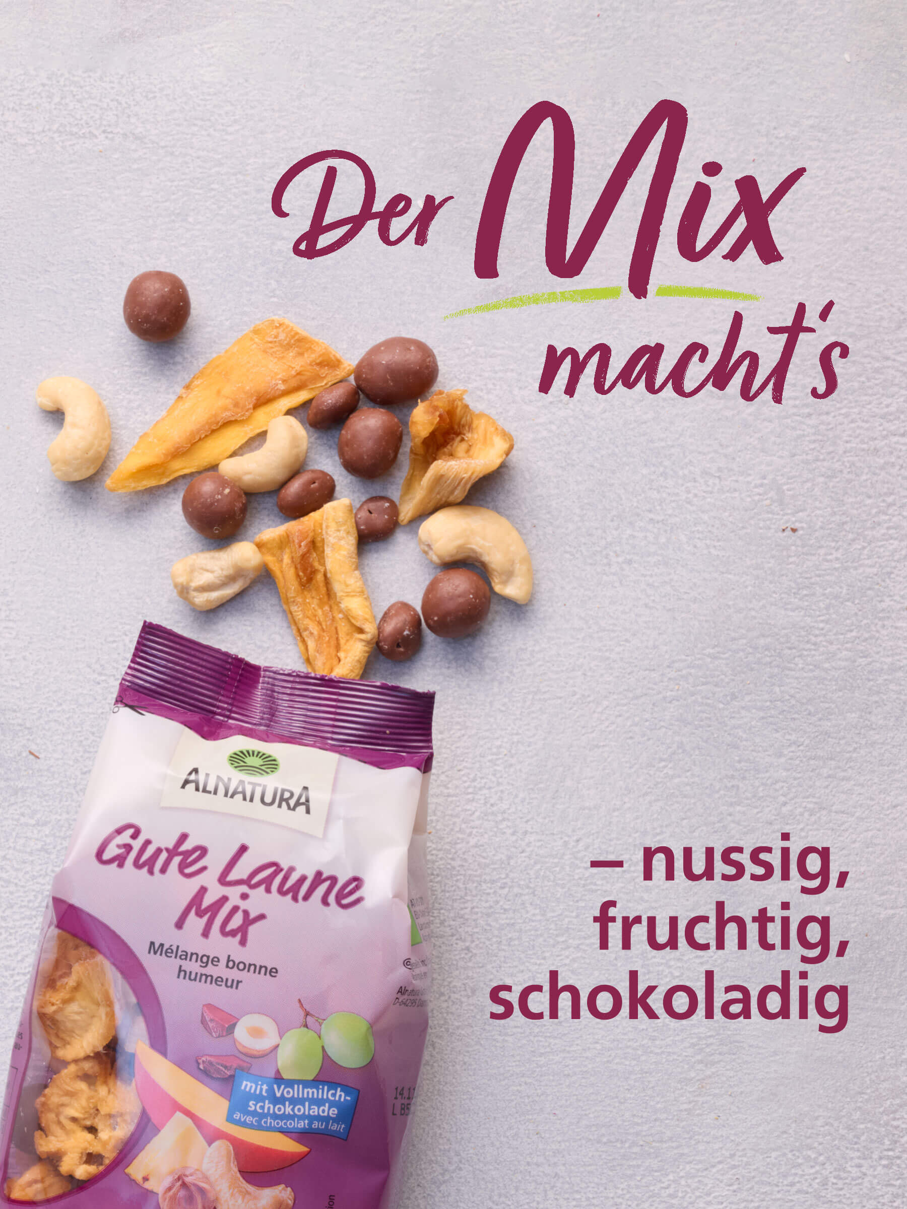 Eine Packung Alnatura Gute Laune Mix liegt auf einem hellen Untergrund, darüber verstreut liegen einige Cashew-Kerne, getrocknete Mangostücke und Schoko-Bällchen. Dazu die Überschrift: "Der Mix macht's - nussig, fruchtig, schokoladig."