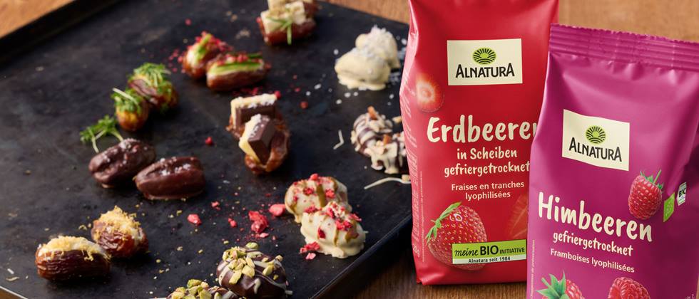 Zwei Alnatura Produkte auf einer Holzunterlage: Erdbeeren gefriergetrocknet und Himbeeren gefriergetrocknet. Daneben ein dunkler, viereckiger Teller, auf dem Datteln mit verschiedenen Toppings drapiert sind. 