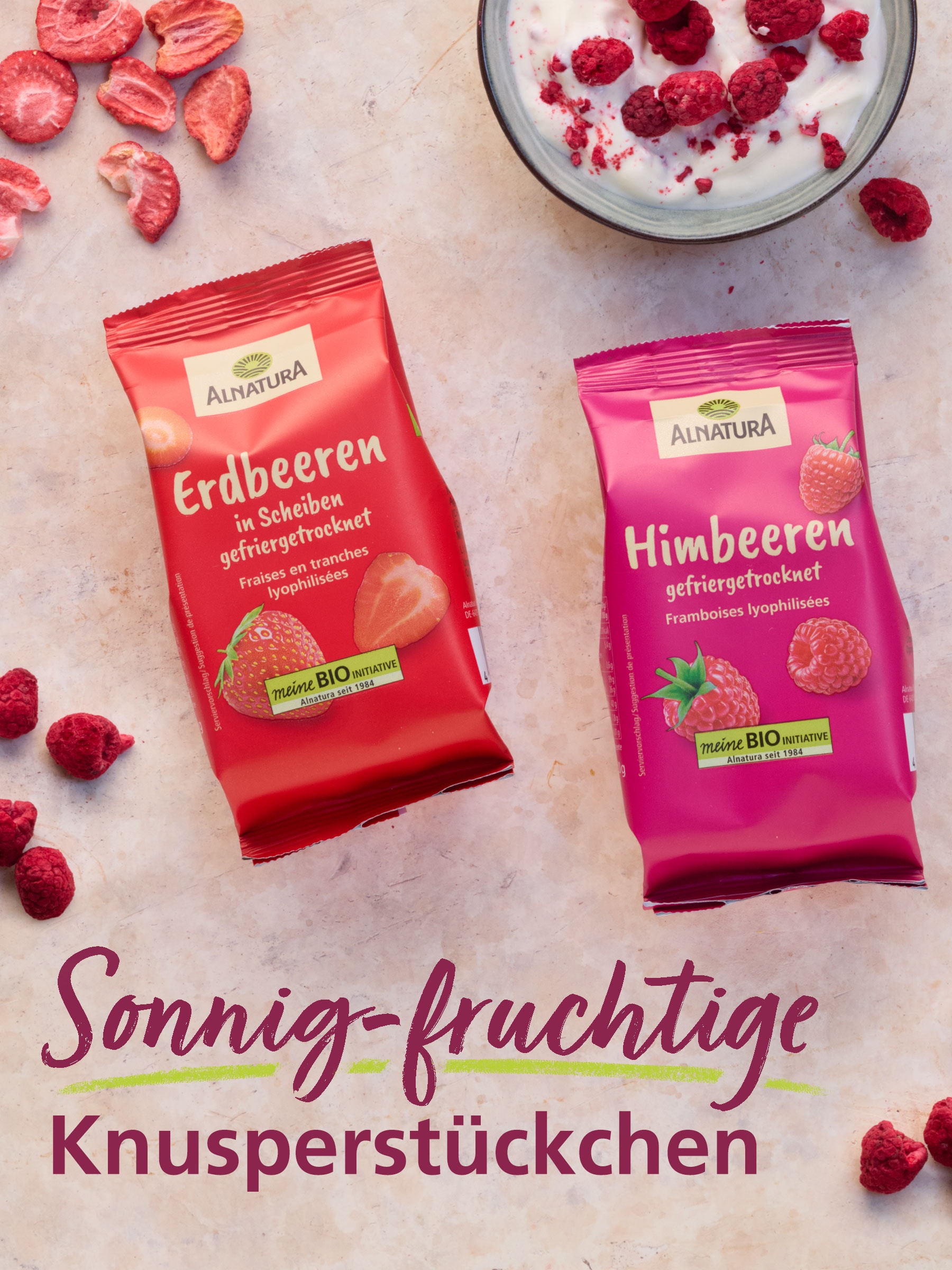 Zwei Alnatura Produkte auf hellem Grund liegend: Gefriergetrocknete Erdbeeren und gefriergetrocknete Himbeeren. Um die Verpackungen sind getrocknete Früchte verteilt. Dazu der Text: "Sonnig-fruchtige Knusperstückchen".
