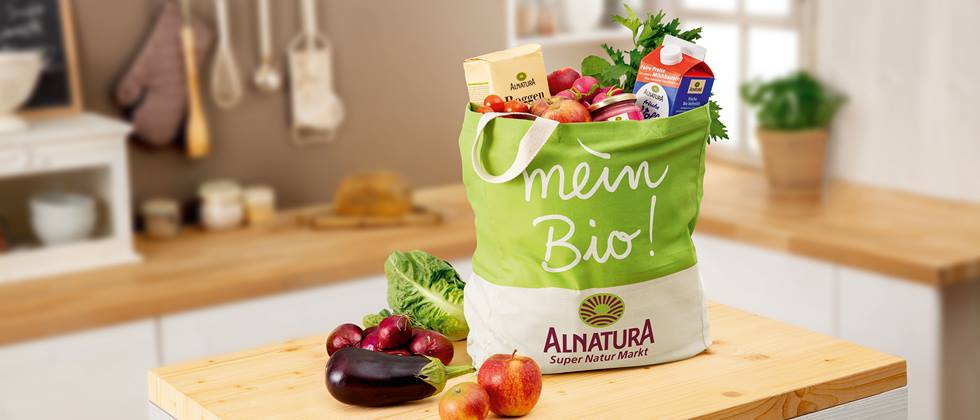 Eine Alnatura-Einkaufstasche prallt gefüllt mit Alnatura Milch, Mehl und Gemüse steht in einer Küche auf einer Kochinsel. Auf dieser liegen noch Äpfel, eine Aubergine, Zwiebeln und ein Romana-Salat. Im Hintergrund ist verschwommen die Küche mit einer weiteren Arbeitsfläche zu sehen.