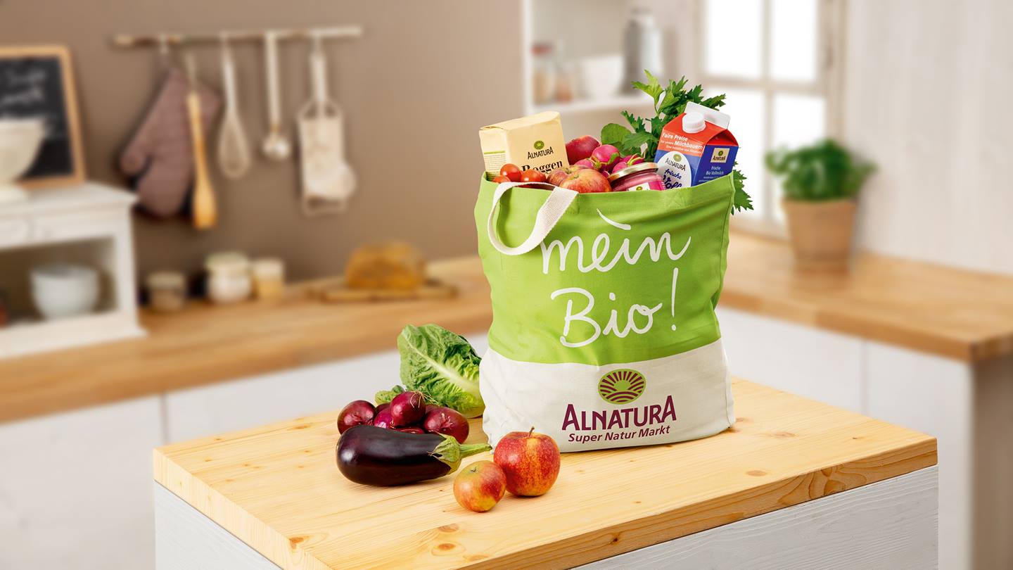 Eine Alnatura-Einkaufstasche prallt gefüllt mit Alnatura Milch, Mehl und Gemüse steht in einer Küche auf einer Kochinsel. Auf dieser liegen noch Äpfel, eine Aubergine, Zwiebeln und ein Romana-Salat. Im Hintergrund ist verschwommen die Küche mit einer weiteren Arbeitsfläche zu sehen.