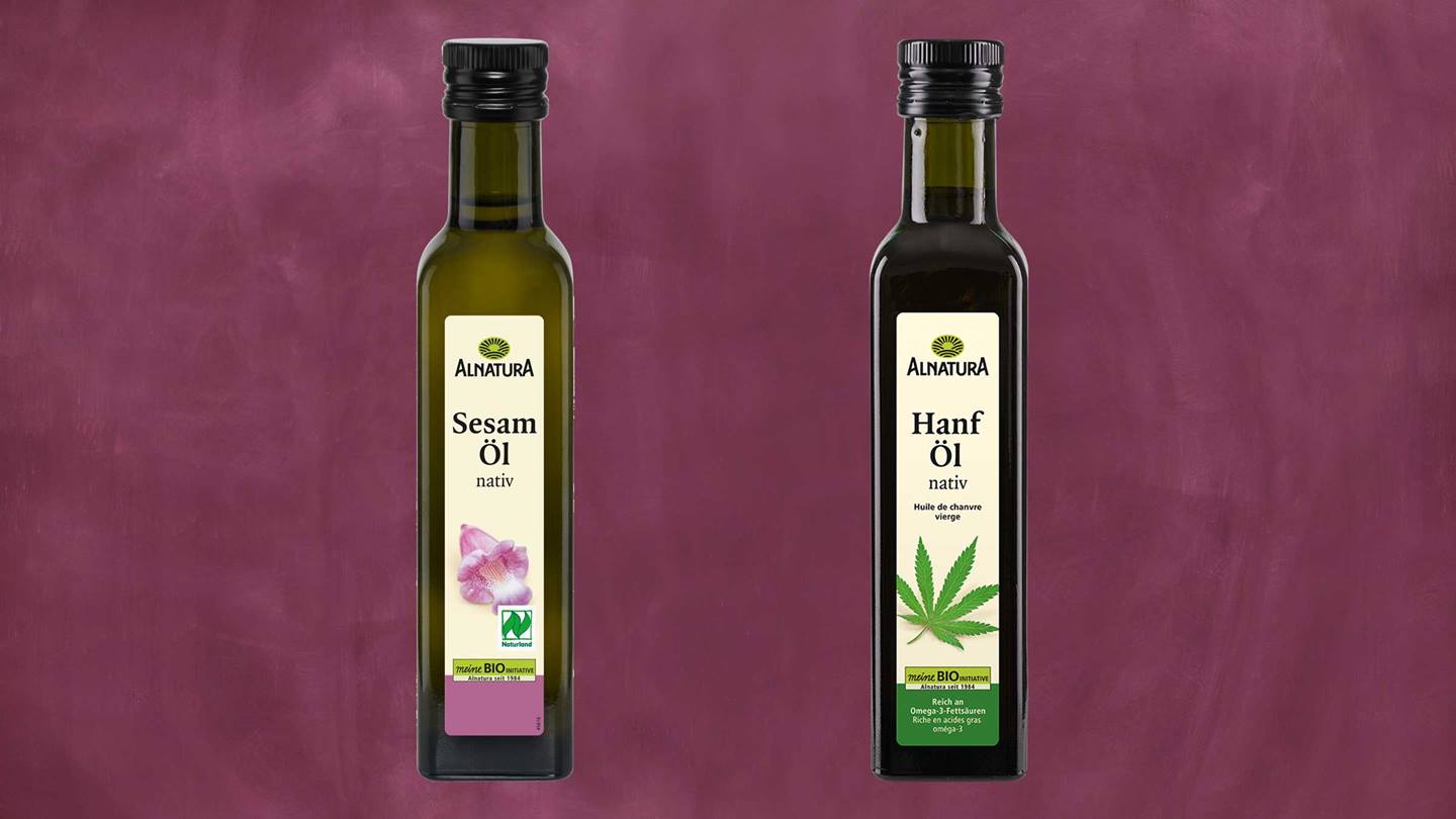 Das Bild zeigt zwei Flaschen natives Öl der Marke Alnatura vor einem weinroten Hintergrund. Links steht eine Flasche Sesamöl, rechts eine Flasche Hanföl. Auf beiden Etiketten ist das Alnatura-Logo, der Hinweis "nativ" und Informationen zur Bio-Qualität abgebildet. Zusätzlich sind kleine Abbildungen der jeweiligen Pflanzen (Sesam und Hanf) zu sehen.