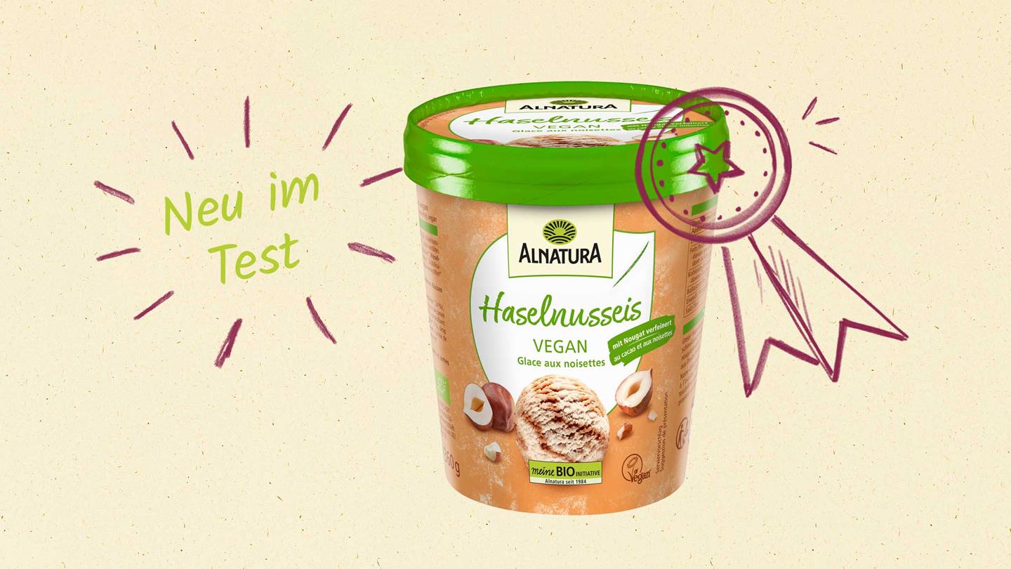 Das Bild zeigt eine Verpackung von veganem Haselnusseis der Marke Alnatura. Die Verpackung ist in beige und grün gehalten und zeigt ein Bild von Haselnusseis mit ganzen Haselnüssen. Rechts neben der Verpackung ist eine handgezeichnete Auszeichnung in Form einer Medaille mit einem Stern zu sehen. Links im Bild steht "Neu im Test" in handschriftlicher Schrift. Insgesamt vermittelt das Bild den Eindruck von einem leckeren und hochwertigen veganen Eisprodukt.