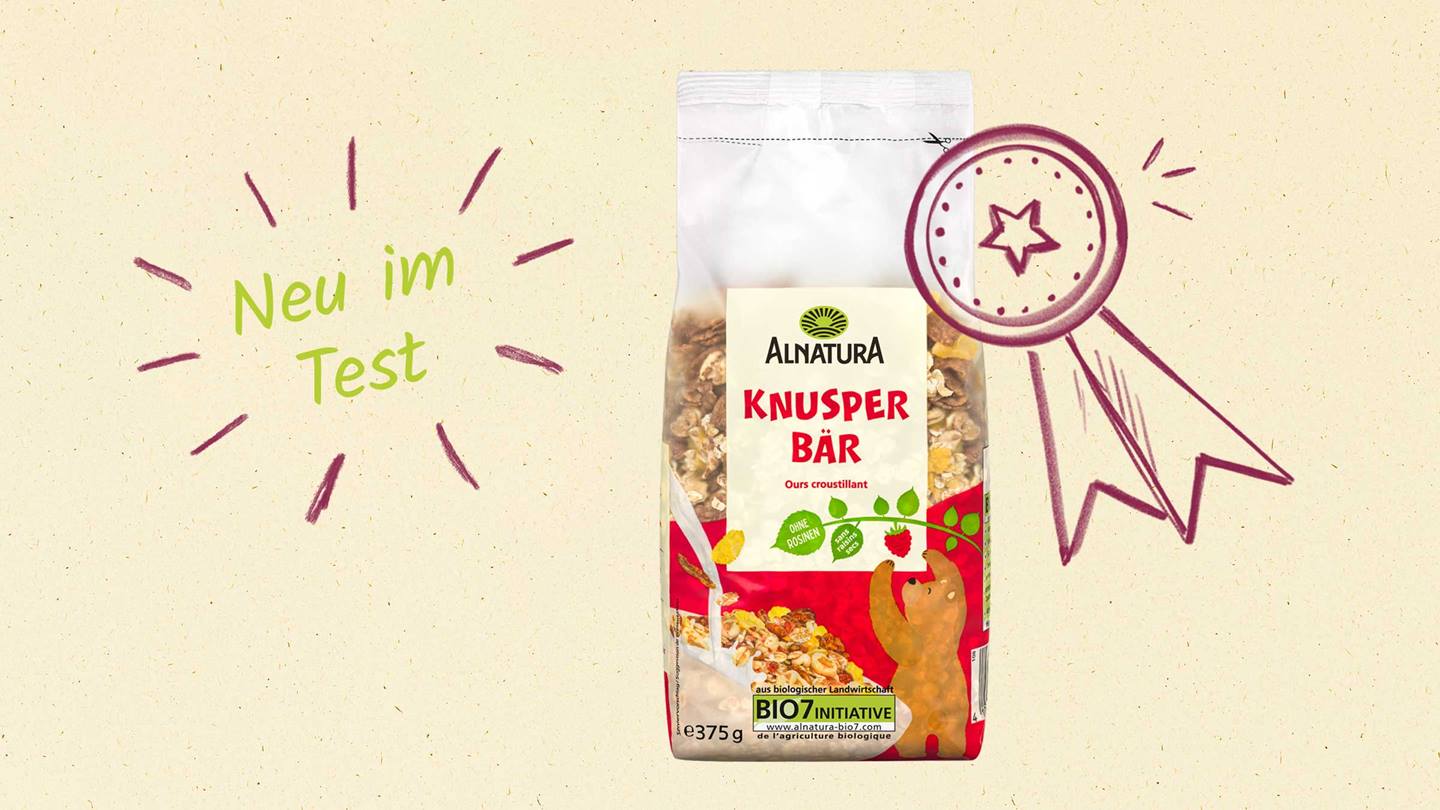Das Bild zeigt eine Packung Alnatura Knusper Bär Müsli. Die Packung ist mit einem handgezeichneten Preisband und dem Text „Neu im Test“ versehen, was auf eine Produktneuheit oder einen Produkttest hinweist. Ein Bärenmotiv ziert die Verpackung. Das Müsli wird als Bio-Produkt beworben, wie der Hinweis auf die BIO7 Initiative verdeutlicht.