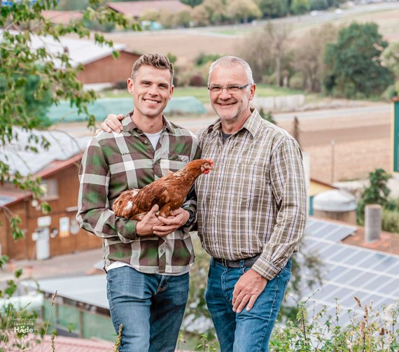 Zwei Männer in karierten Hemden stehen vor einer ländlichen Kulisse, einer hält ein braunes Huhn.
