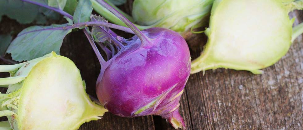 Das Bild zeigt mehrere Kohlrabi auf einem hölzernen Untergrund. Ein Kohlrabi ist violett und im Vordergrund, während zwei weitere hellgrüne Kohlrabi im Hintergrund liegen. Ein Teil jedes der hellgrünen Kohlrabi ist durchgeschnitten, um das Innere zu zeigen. Die Kohlrabi sind frisch und sehen appetitlich aus. Das Bild ist einfach, aber ansprechend und betont die Farben und Texturen der Kohlrabi.