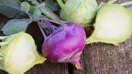 Das Bild zeigt mehrere Kohlrabi auf einem hölzernen Untergrund. Ein Kohlrabi ist violett und im Vordergrund, während zwei weitere hellgrüne Kohlrabi im Hintergrund liegen. Ein Teil jedes der hellgrünen Kohlrabi ist durchgeschnitten, um das Innere zu zeigen. Die Kohlrabi sind frisch und sehen appetitlich aus. Das Bild ist einfach, aber ansprechend und betont die Farben und Texturen der Kohlrabi.