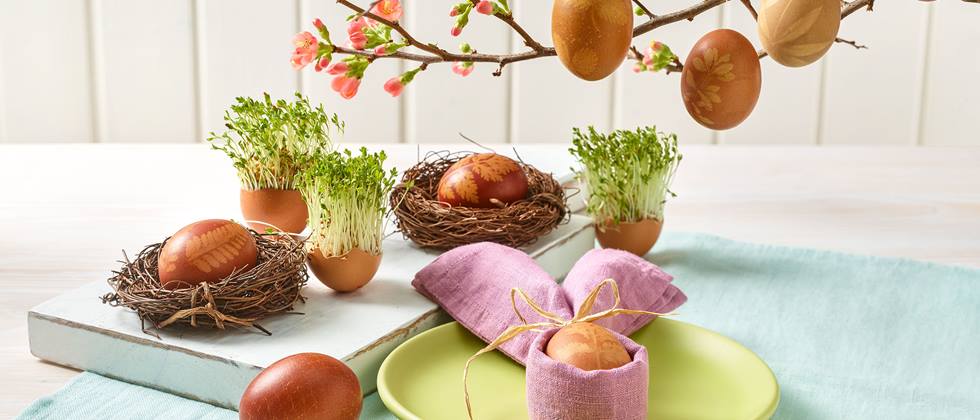 Das Bild zeigt eine festliche Osterdekoration. Mehrere rotbraune Ostereier, mit Pflanzenmotiven verziert, sind in kleinen Nestern aus Zweigen arrangiert.  Weitere bemalte Eier hängen an einem blühenden Zweig.  Kresse in Eierschalen ergänzt die Dekoration.  Im Vordergrund steht ein Tischgedeck mit einem weiteren verzierten Ei, das in einem lilafarbenen Stofftuch liegt. Das Gesamtbild wirkt frühlingshaft, natürlich und elegant.