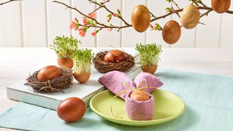 Das Bild zeigt eine festliche Osterdekoration. Mehrere rotbraune Ostereier, mit Pflanzenmotiven verziert, sind in kleinen Nestern aus Zweigen arrangiert.  Weitere bemalte Eier hängen an einem blühenden Zweig.  Kresse in Eierschalen ergänzt die Dekoration.  Im Vordergrund steht ein Tischgedeck mit einem weiteren verzierten Ei, das in einem lilafarbenen Stofftuch liegt. Das Gesamtbild wirkt frühlingshaft, natürlich und elegant.