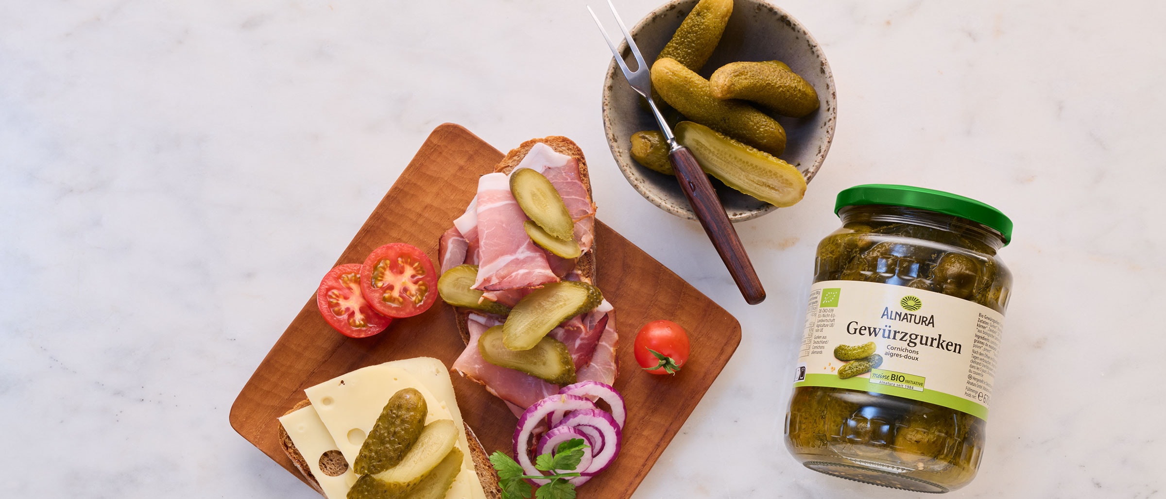 Brett mit Brot, Käse, Schinken, Gewürzgurken, Tomaten und Zwiebeln neben einem Glas Gewürzgurken und einer Schale mit mehreren Gewürzgurken und einer Gabel