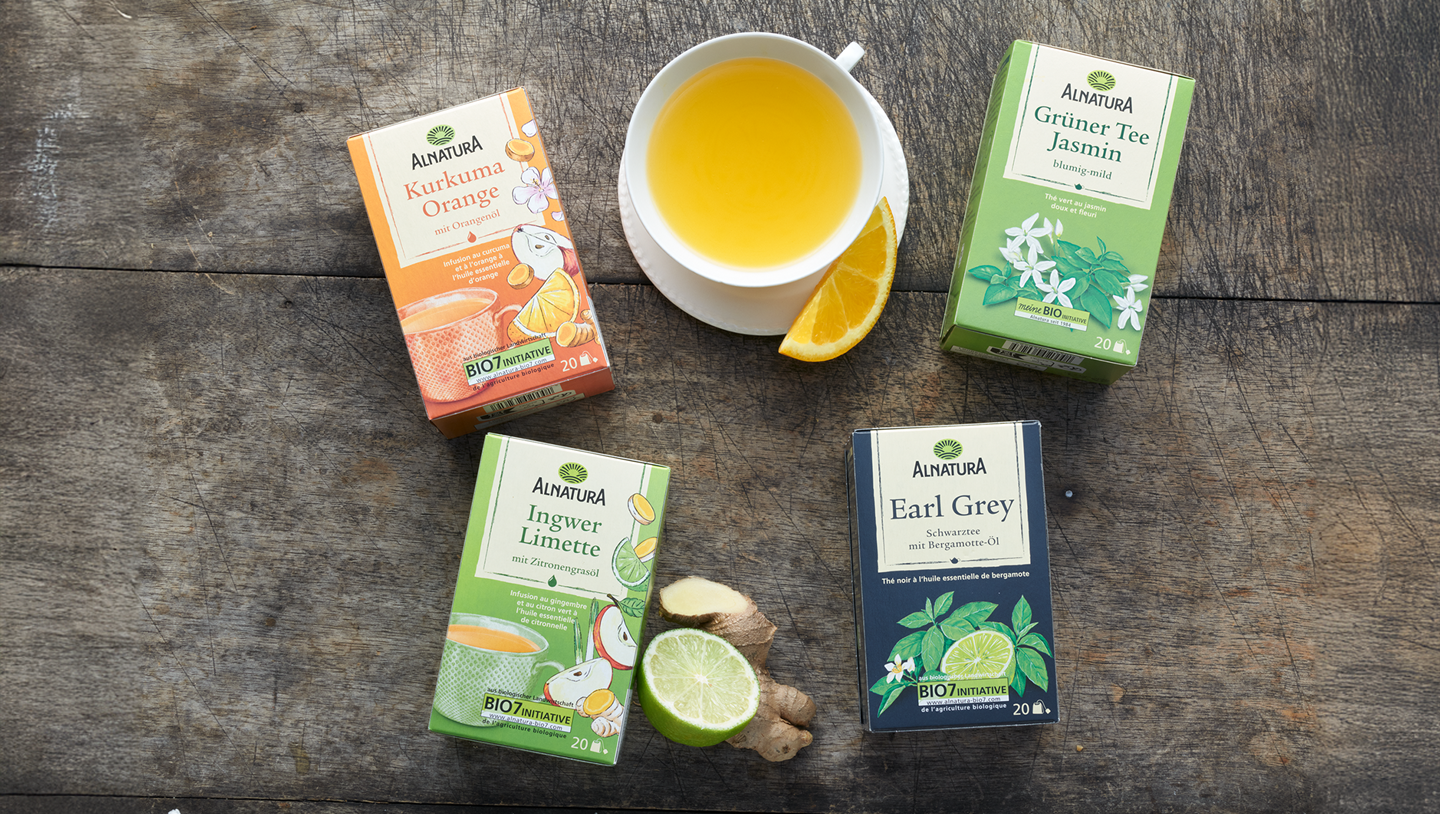 Verschiedene Alnatura Tee-Packungen liegen auf einem dunklen Holzuntergrund, daneben eine Tasse mit hellem Tee und einem Stück Orange