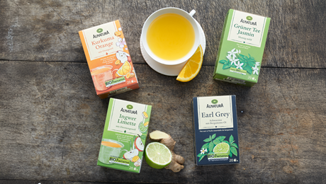 Verschiedene Alnatura Tee-Packungen liegen auf einem dunklen Holzuntergrund, daneben eine Tasse mit hellem Tee und einem Stück Orange