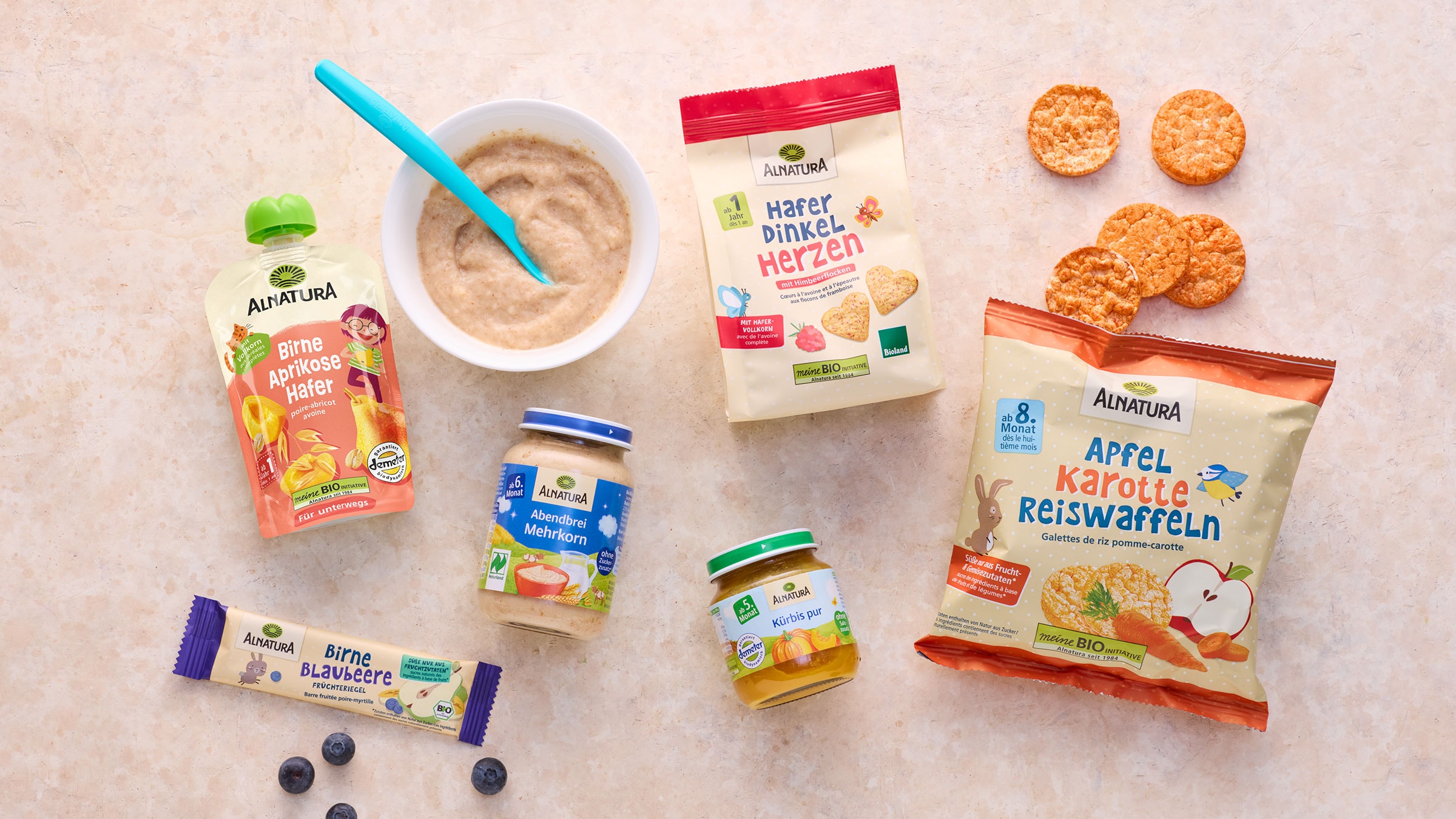 Alnatura Babykostprodukte, darunter Babybrei, Fruchtmus, Kekse und Reiswaffeln, liegen auf einem beigefarbenen Hintergrund, daneben einige blaue Beeren.