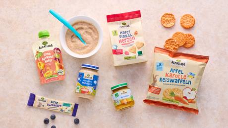 Alnatura Babykostprodukte, darunter Babybrei, Fruchtmus, Kekse und Reiswaffeln, liegen auf einem beigefarbenen Hintergrund, daneben einige blaue Beeren.