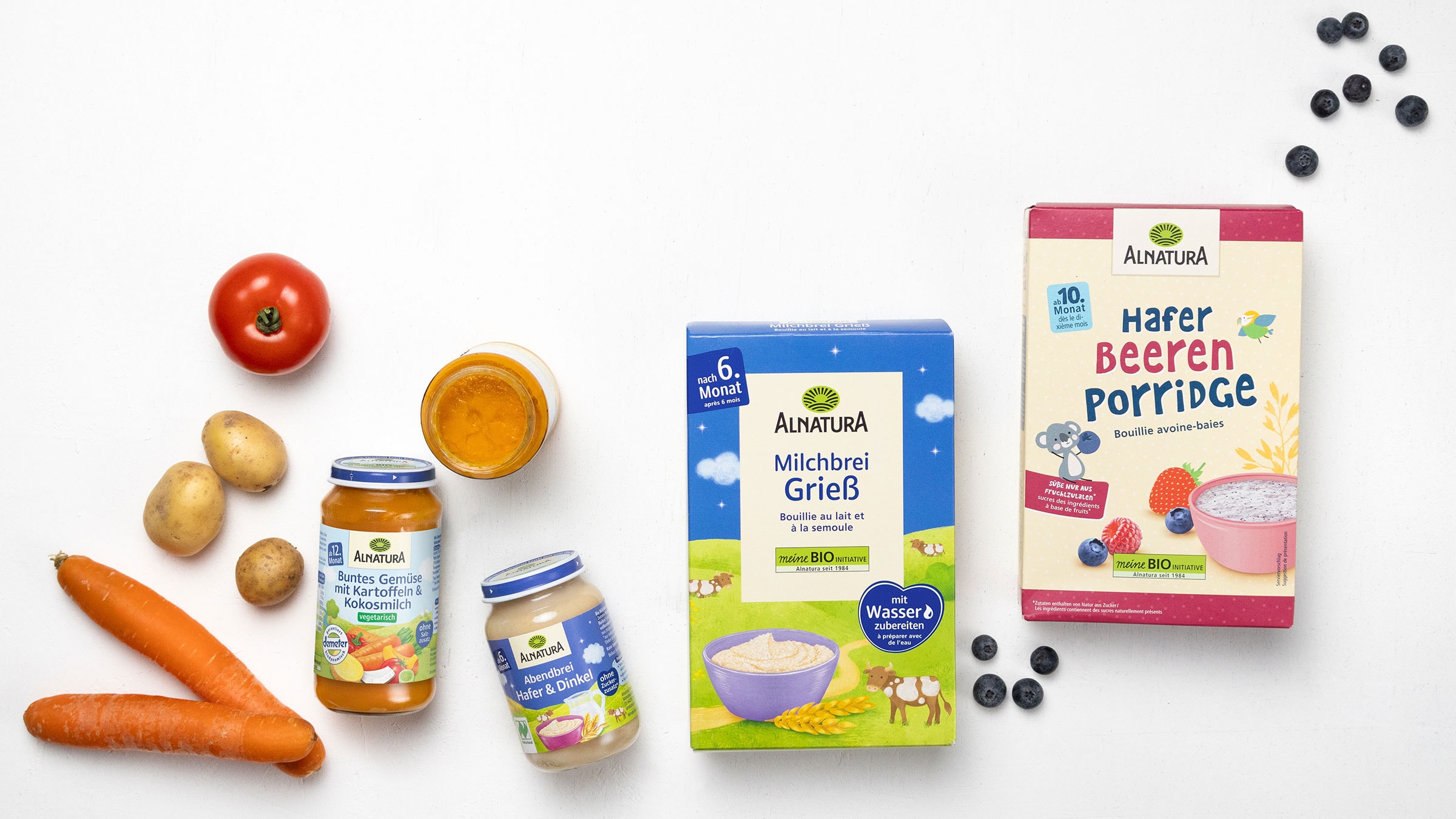 Eine Auswahl an Alnatura-Baby-Produkten sowie frisches Gemüse und Beeren: Drei Gläser mit verschiedenen Breisorten: "Buntes Gemüse mit Kartoffeln & Kokosmilch", "Abendbrei Hafer & Dinkel" und ein geöffnetes Glas mit einem orangefarbenen Brei, zwei Packungen Getreidebrei: "Milchbrei Grieß" (ab dem 6. Monat) und "Hafer-Beeren-Porridge" um die Produkte herum liegen frische Karotten, Kartoffeln, eine Tomate und Blaubeeren.