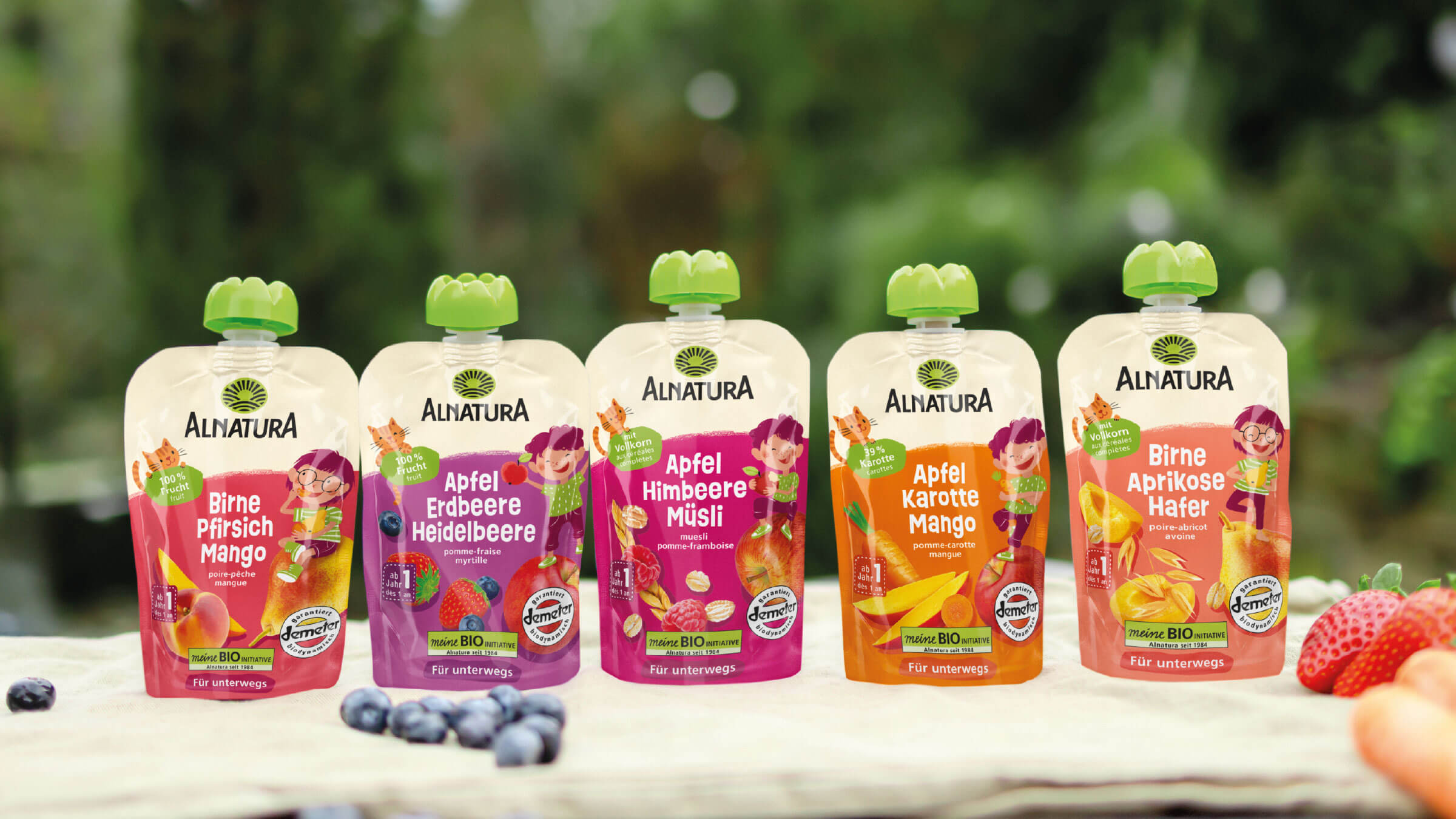 Fünf bunte Alnatura-Fruchtbeutel für unterwegs, aufgereiht auf einer natürlichen Holzfläche mit unscharfem Grün im Hintergrund. Die Beutel zeigen verschiedene Geschmackssorten: Birne-Pfirsich-Mango, Apfel-Erdbeere-Heidelbeere, Apfel-Himbeere-Müsli, Apfel-Karotte-Mango und Birne-Aprikose-Hafer.