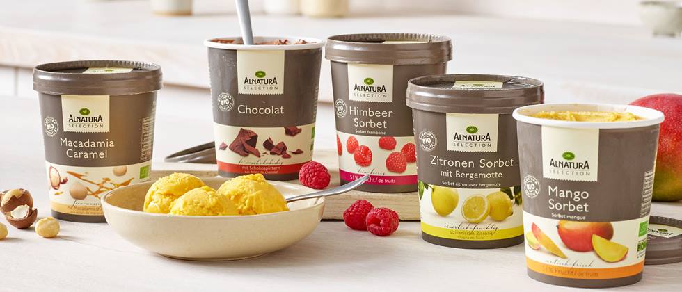 Mehrere Becher mit verschiedenen Eissorten von Alnatura stehen auf einem Tisch, daneben eine Schüssel mit Eis, Himbeeren und Zitronen.