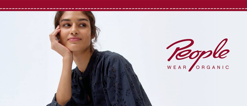 Eine brünette Dame in einer dunkelblauen Stoffbluse stützt den Kopf auf rechten Arm und blickt an der Kamera vorbei. Der Hintergrund ist gräulich und es ist der Schriftzug der Marke People Wear Organic zu lesen,
