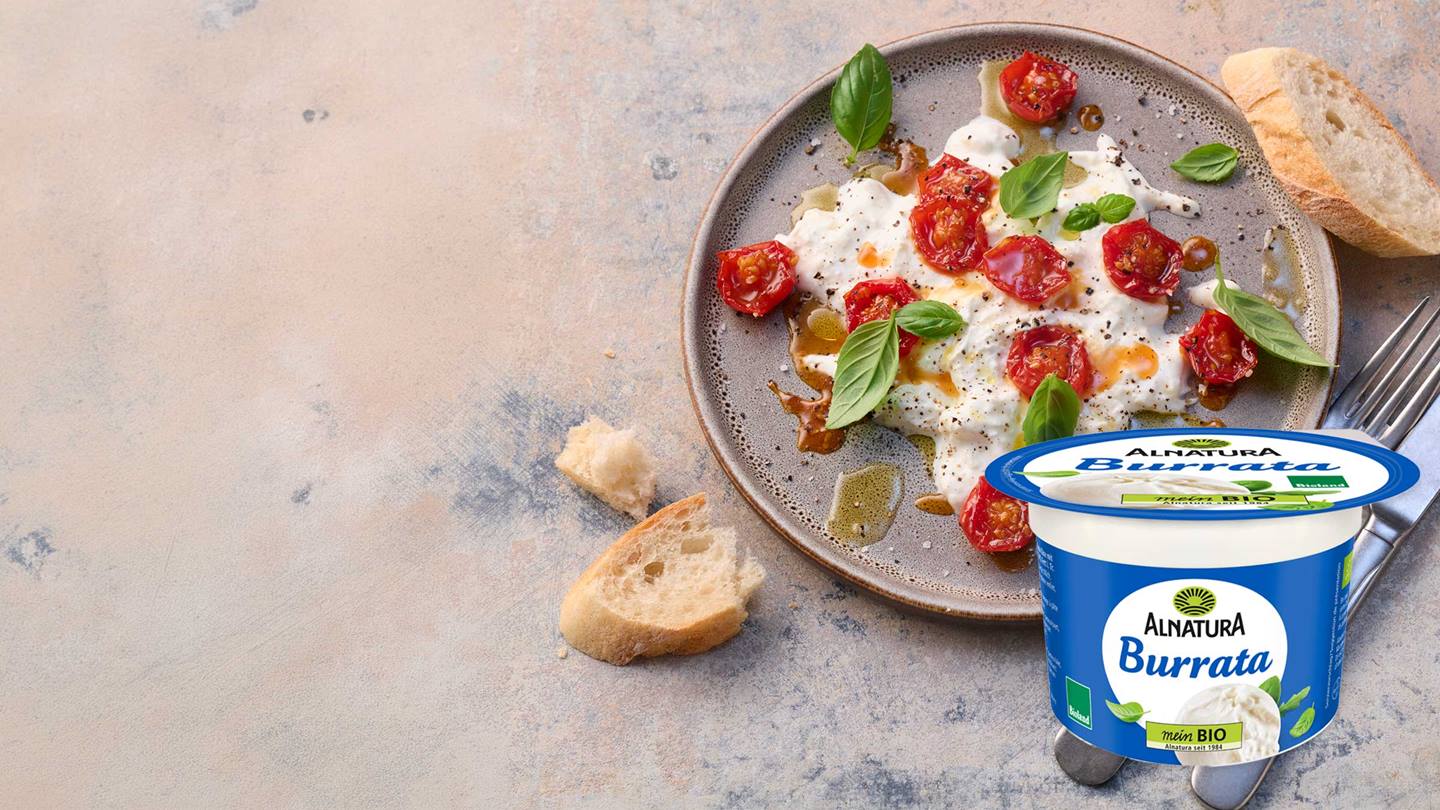 Ein Teller mit einer Burrata, die mit Kirschtomaten und Basilikum garniert ist, daneben eine Packung Alnatura Burrata und Baguette.