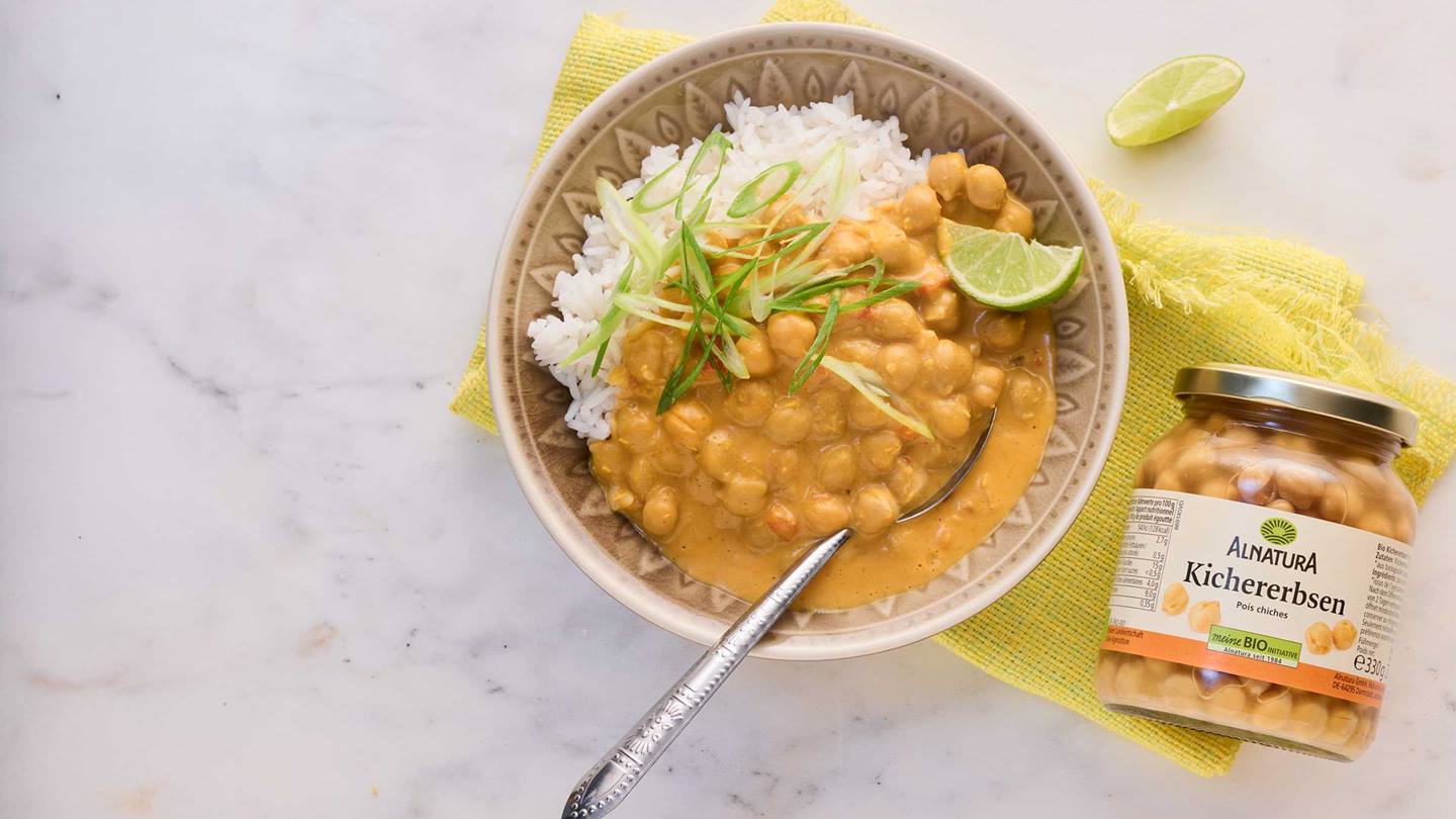 Eine Schale mit Kichererbsencurry und Reis, garniert mit Lauchzwiebeln und Limette, steht auf einer gelben Stoffunterlage neben einem Glas Alnatura Kichererbsen.