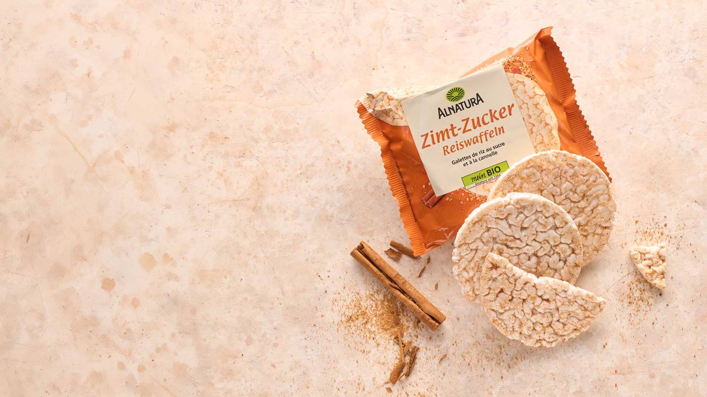 Eine Packung Alnatura Zimt-Zucker Reiswaffeln liegt neben drei Reiswaffeln und Zimtstangen auf einem hellbraunen Untergrund.