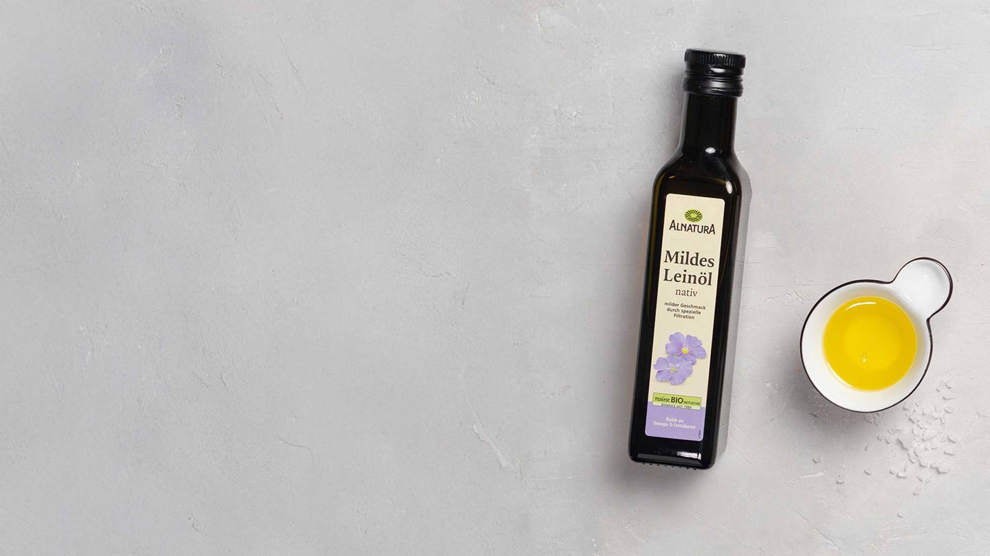 Das Bild zeigt eine Flasche Alnatura Mildes Leinöl nativ auf einem hellen, grauen Untergrund. Neben der Flasche befindet sich eine kleine Schale mit Leinöl und etwas grobes Salz.  Das Bild ist einfach, sauber und wirkt werblich. Der Fokus liegt auf dem Produkt und dessen Eigenschaften (milder Geschmack, reich an Omega-3-Fettsäuren).
