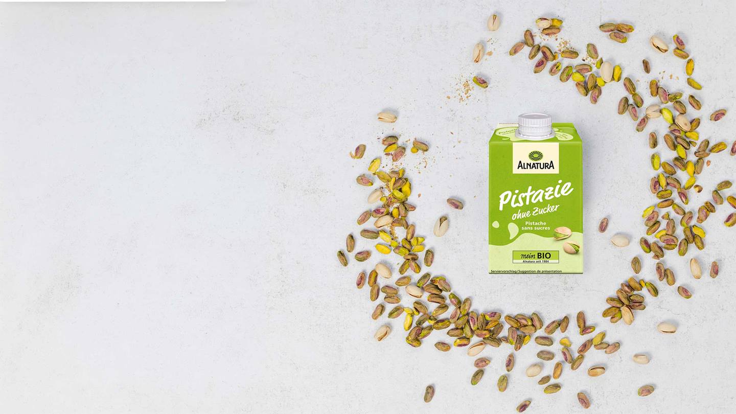 Ein Getränkekarton mit der Aufschrift "Alnatura Pistazie ohne Zucker" steht neben einer Streuung von Pistazien auf einem hellen Untergrund.