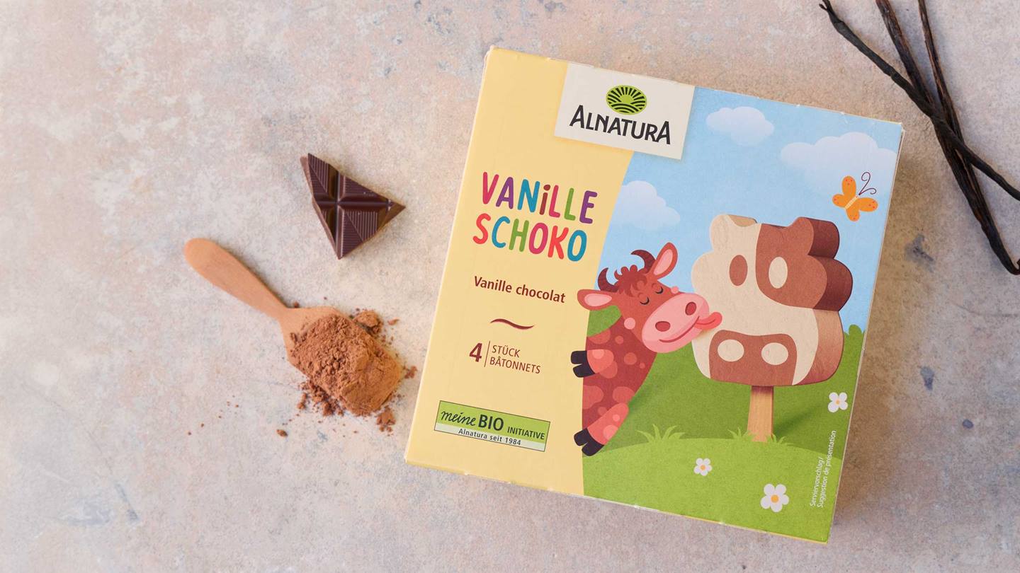 Das Bild zeigt eine Packung Alnatura Vanille Schoko-Eis. Auf der Verpackung ist eine niedliche Kuh abgebildet, die an einem Stieleis leckt, das aus Vanille und Schokolade besteht. Neben der Packung liegen ein Stück dunkle Schokolade und ein Holzlöffel mit Kakaopulver. Im Hintergrund sind Vanilleschoten zu sehen. Das Gesamtbild vermittelt einen Eindruck von Genuss und Natürlichkeit.