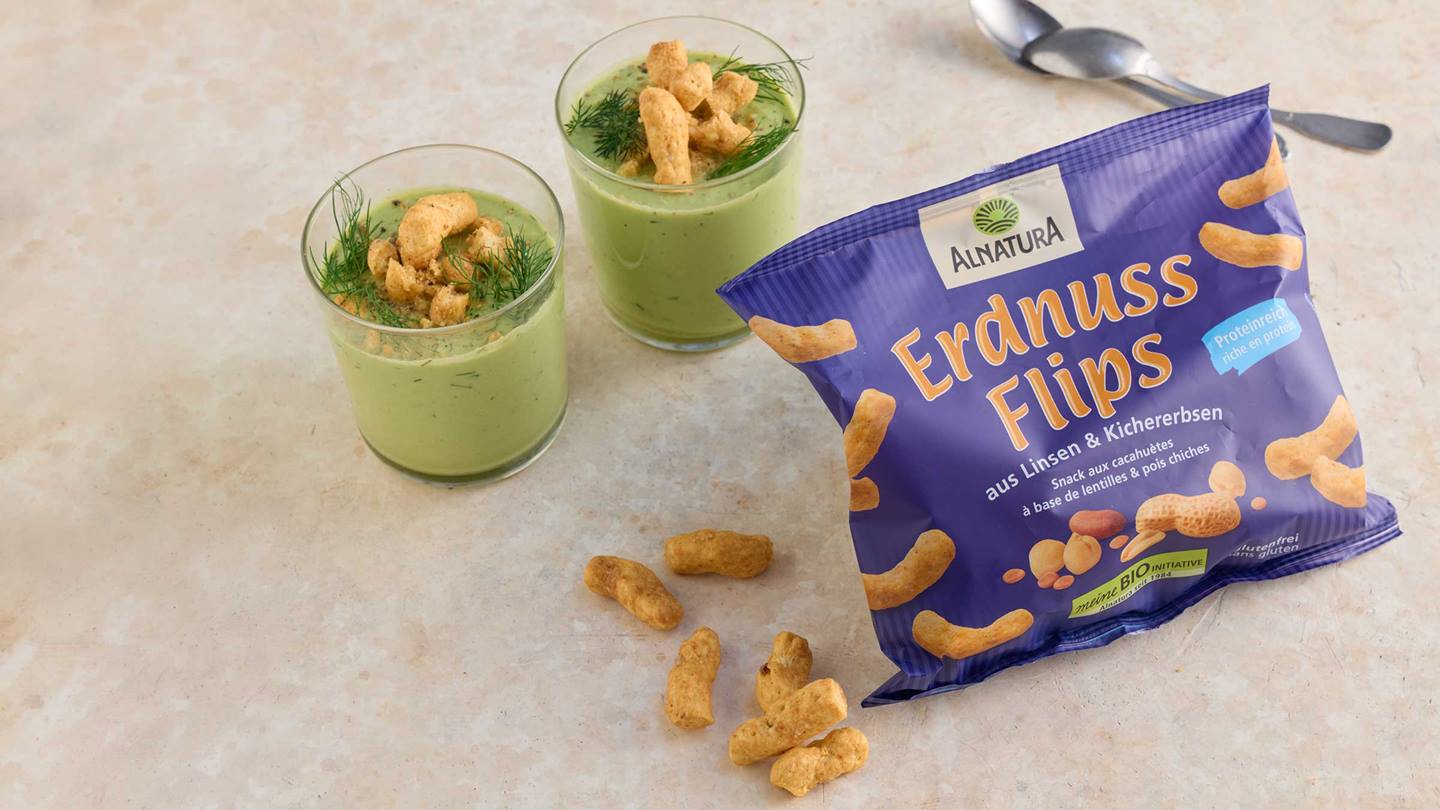 Eine Tüte Alnatura Erdnussflips Linsen & Kichererbsen neben zwei Gläsern mit einer grünen Suppe, die mit Dill und Erdnussflips dekoriert sind.