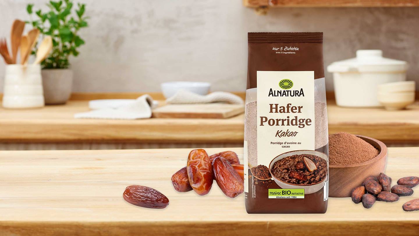 Das Bild zeigt eine Packung Alnatura Hafer Porridge Kakao auf einem Holztisch, umgeben von Datteln, Kakaopulver und Kakaobohnen.