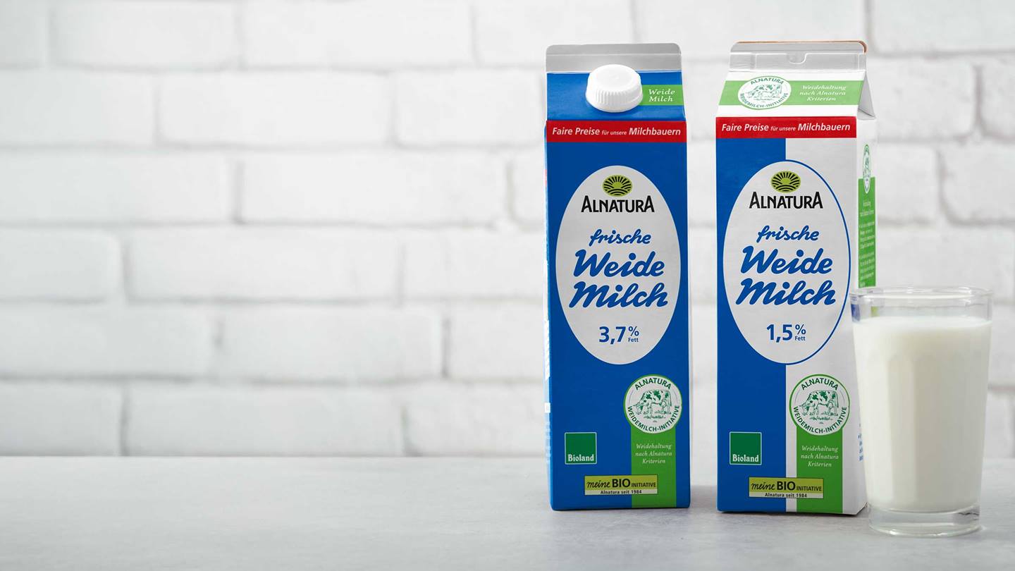 Zwei Alnatura Weidemilch-Packungen mit unterschiedlichem Fettgehalt stehen neben einem Glas Milch vor einer weißen Ziegelwand.