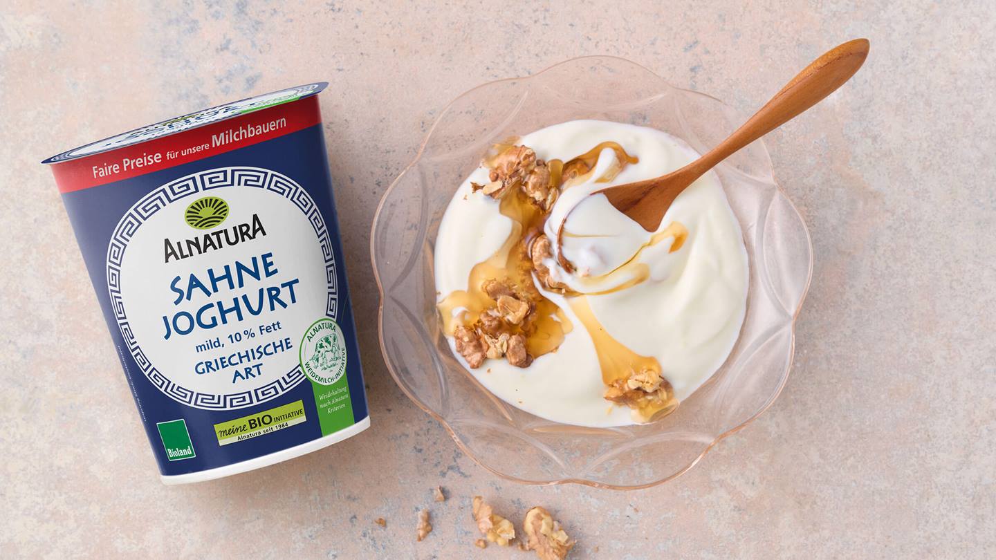 Das Bild zeigt eine Schale mit griechischem Joghurt von Alnatura. Der Joghurt ist mit Honig und Walnüssen verfeinert. Neben der Schale steht ein Becher mit dem gleichen Joghurt.  Die Szene ist hell und freundlich gestaltet und vermittelt ein Gefühl von Natürlichkeit und Frische.