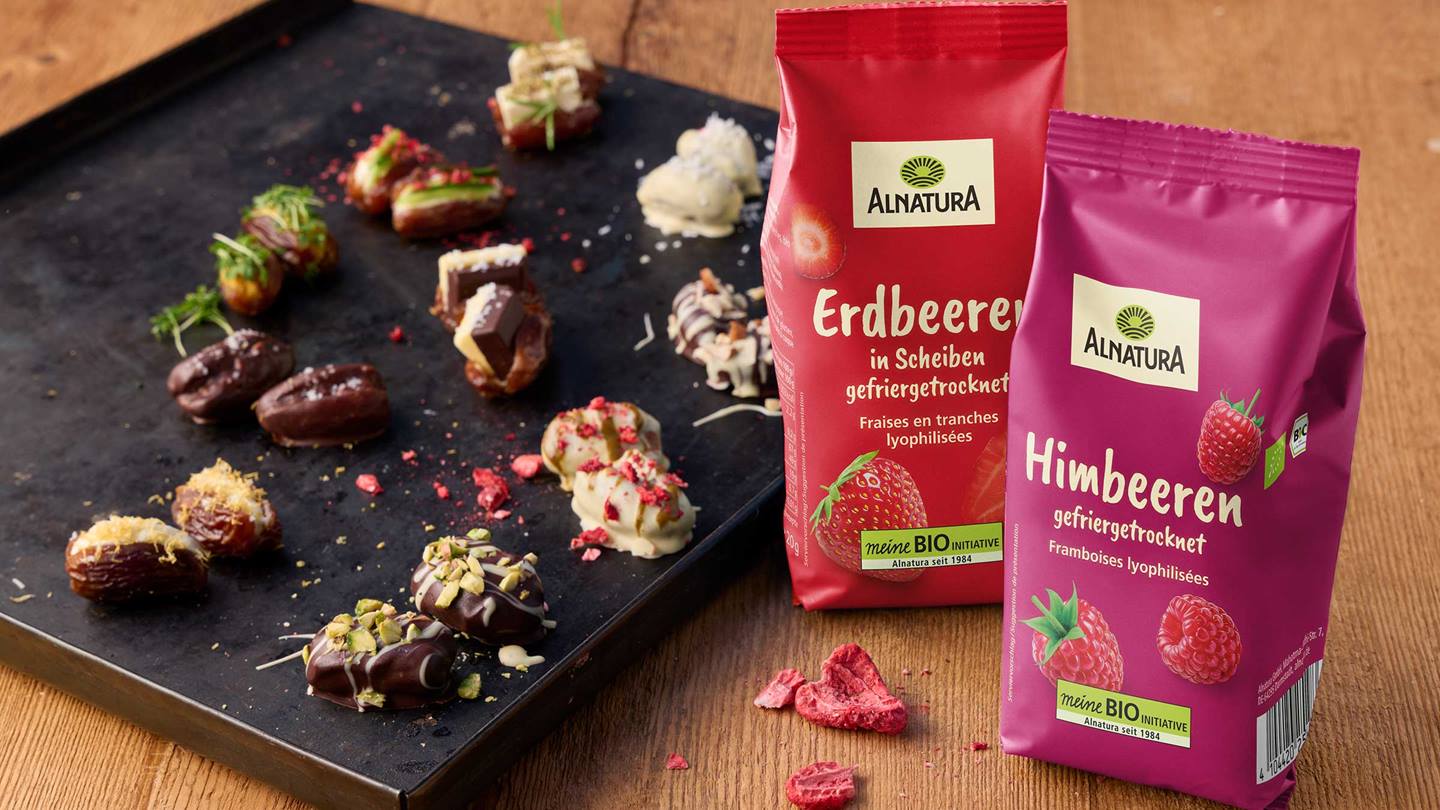 Das Bild zeigt verschiedene Datteln, die mit Schokolade überzogen und mit verschiedenen Toppings (Pistazien, getrocknete Erdbeeren und Himbeeren) dekoriert sind. Im Vordergrund stehen zwei Packungen gefriergetrockneter Erdbeeren und Himbeeren der Marke Alnatura. Das Gesamtbild wirkt appetitlich und stellt eine ansprechende Kombination aus süßen Snacks und Bio-Produkten dar.