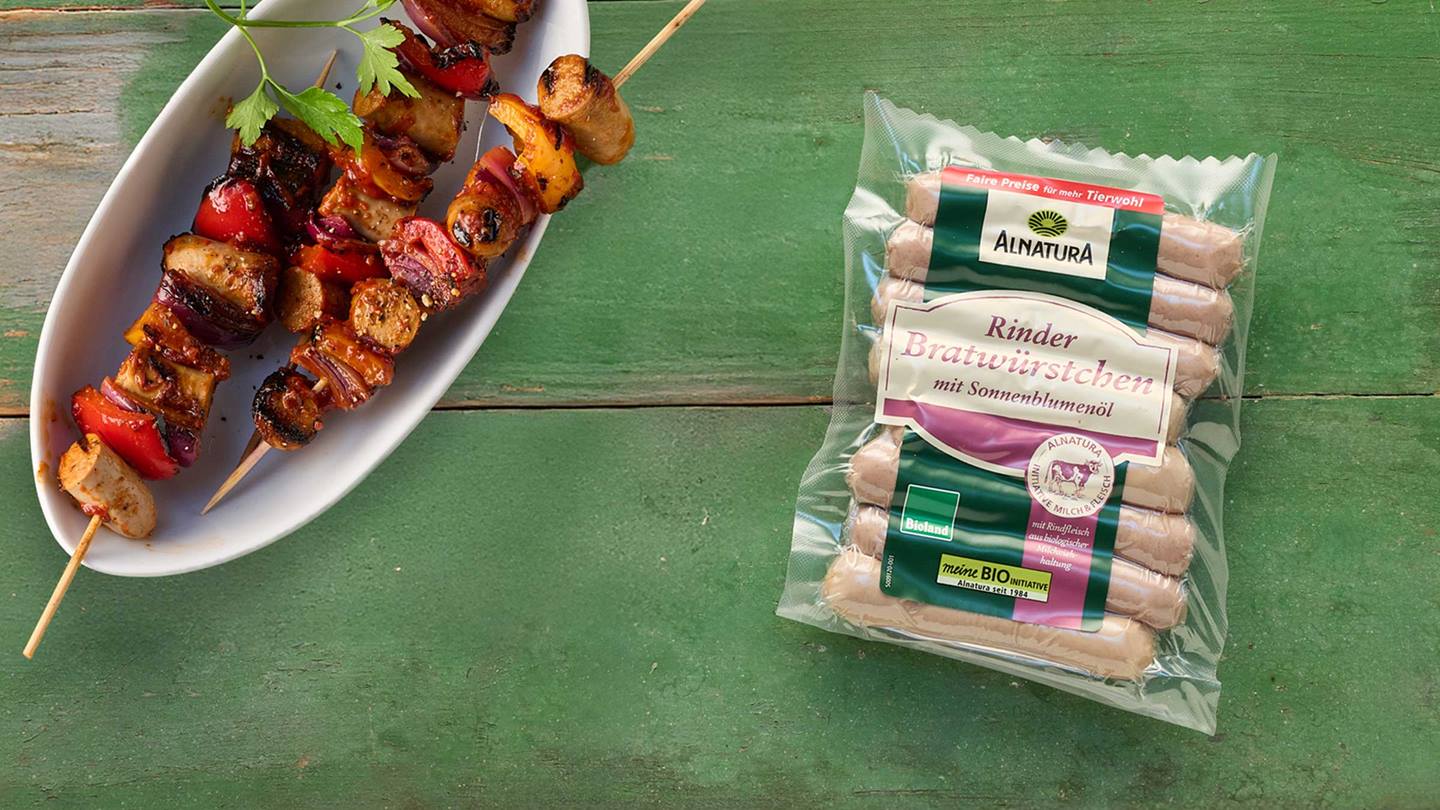 Das Bild zeigt zwei Spieße mit Rindfleischbratwürstchen, Paprika, Zwiebeln und anderen Gemüsesorten, die auf einem weißen Teller auf einem grünen Holzhintergrund angerichtet sind. Neben dem Teller liegt eine Verpackung mit Alnatura Rindfleischbratwürstchen aus Bioland-Produktion. Das Gesamtbild wirkt appetitlich und suggeriert ein gesundes und leckeres Gericht.