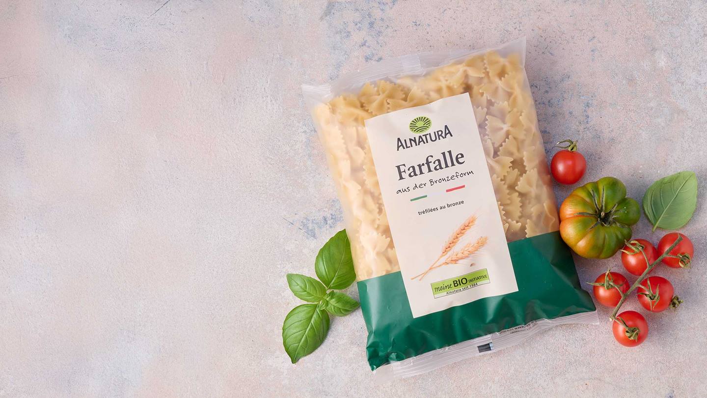 Das Bild zeigt eine Packung Alnatura Bio Farfalle aus Bronzeform. Die Packung liegt auf einer hellen, leicht strukturierten Oberfläche. Neben der Pasta liegen einige Kirschtomaten und ein Zweig Basilikum.  Das Bild wirkt appetitanregend und vermittelt Frische und Natürlichkeit.  Es ist offensichtlich eine Produktfotografie, die die Pasta und ihre Zutaten in Szene setzt.