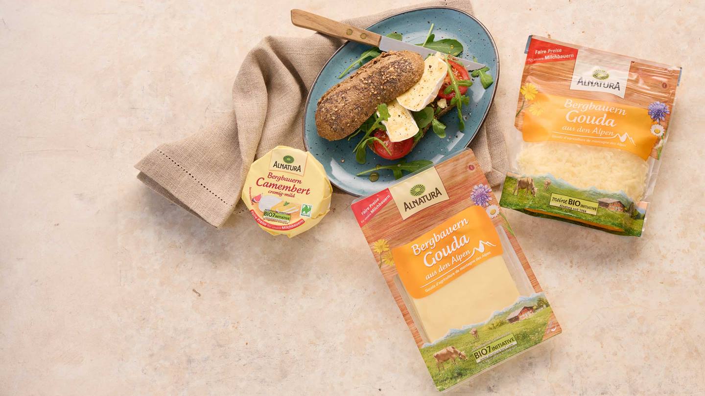 Das Bild zeigt ein Brötchen mit Camembert, Tomaten und Rucola auf einem Teller.  Neben dem Teller liegen zwei Verpackungen Alnatura Bergbauer Käse: eine mit geriebenem Gouda und eine mit einer Scheibe Gouda.  Eine weitere Verpackung mit Camembert ist ebenfalls abgebildet. Das Gesamtbild suggeriert eine ansprechende, einfache und wohlschmeckende Mahlzeit mit Bio-Produkten.