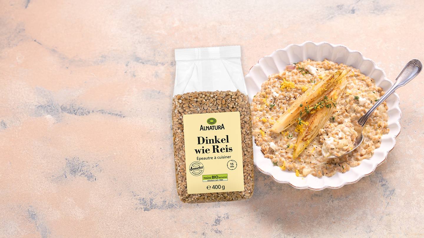 Das Bild zeigt eine Packung Alnatura Dinkel wie Reis (400g) neben einer weißen Schüssel mit einem Gericht aus diesem Dinkel, Chicorée und einer cremigen Sauce. Das Essen scheint mit Thymian und Zitronenschale garniert zu sein. Der Hintergrund ist ein hellbeiger Untergrund.  Das Bild wirkt appetitlich und werbewirksam und präsentiert ein fertiges Gericht, das mit dem Produkt zubereitet wurde.