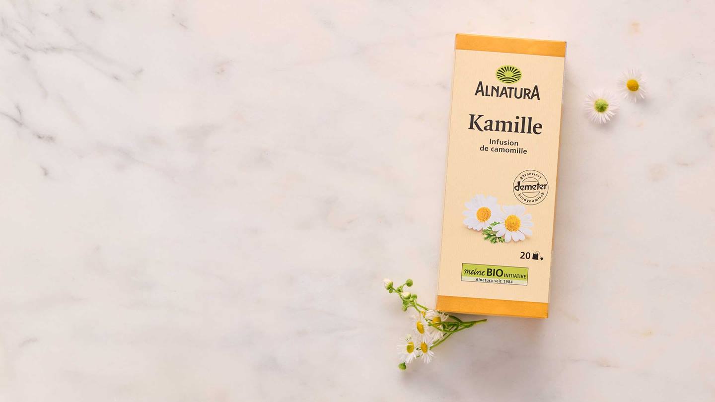 Das Bild zeigt eine Packung Kamillentee von Alnatura, die auf einer hellen, marmorierten Oberfläche liegt. Neben der Packung liegen einige frische Kamillenblüten. Die Packung selbst ist in hellen Farben gehalten und trägt das Alnatura-Logo sowie die Aufschrift "Kamille" und weitere Informationen zum Produkt.