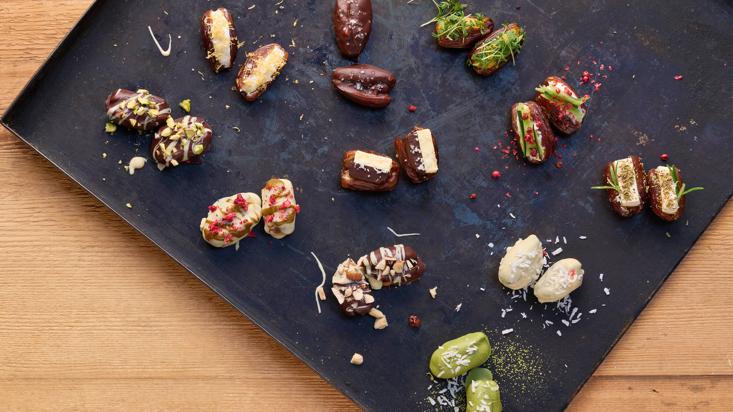 Auf einem Holzuntergrund liegt ein schwarzes Backckblech auf dem viele Datteln liegen, die verschieden verfeinert sind. Zum Beispiel mit  Mandelnstücken betreut, umhüllt mit Matchapulver und weißer Schokolade, gefüllt mit Ricotta oder dunkler Schokolade.