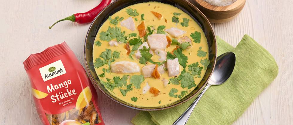 Eine dunkle Porzellan-Schüssel mit einer gelben Kokos-Suppe, einem Curry steht auf einem hellen Holzuntergrund. In dem Curry sind einige Stücke Kabeljau, getrocknete Mangostücke und frischer Koriander. Neben dem Curry steht eine Holzschüssel mit gekochtem Reis und es liegen eine rote Chilischote, eine Packung Alnatura Mango Stücke, ein Löffel und eine grüne Stoffserviette daneben.