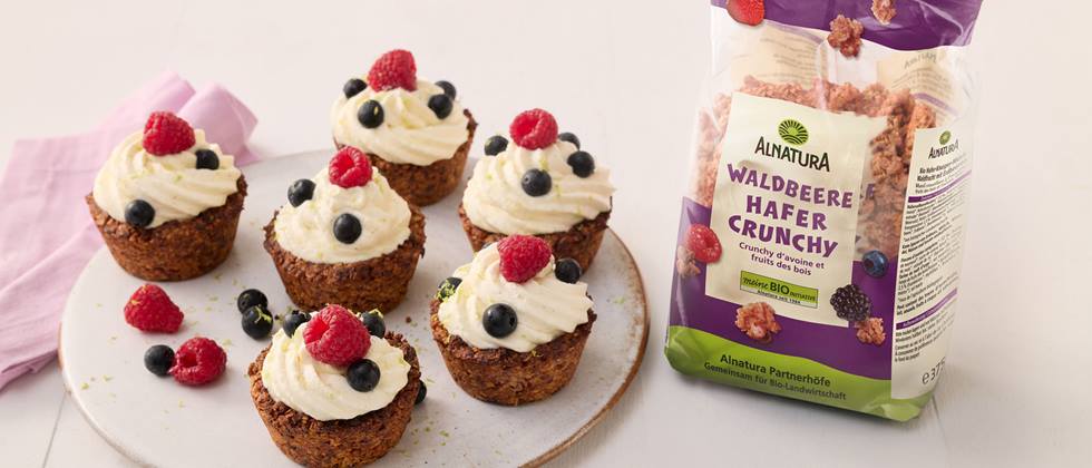 Auf einem weißen Porzellanteller stehen sechs Törtchen in Muffinform aus Müsli hergestellt. Die Törtchen haben eine braune Farbe und ein Sahnehäubchen, garniert mit einer frischen Himbeere und frischen Heidelbeeren. Im Hintergrund ist eine violette Stoffserviette. Neben dem Teller steht eine Packung Alnatura Waldbeere-Hafer-Crunchy