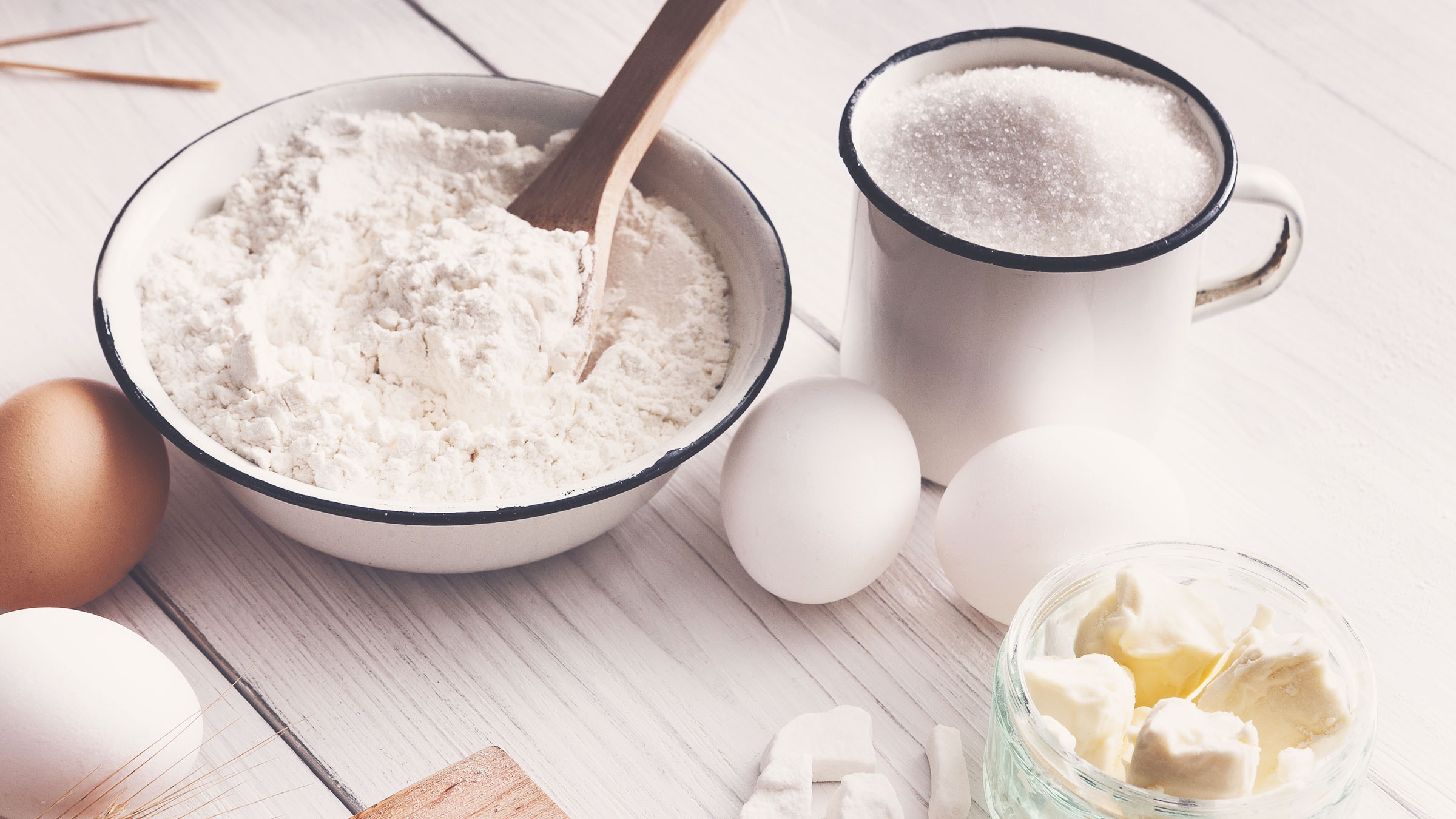 Auf hellem Holzuntergrund steht eine Schüssel mit Mehl, in dieser ein Holzlöffel steckt, eine Tasse Zucker, eine kleine Glasschale mit Butter. Daneben liegen zwei weiße und ein braunes Ei.