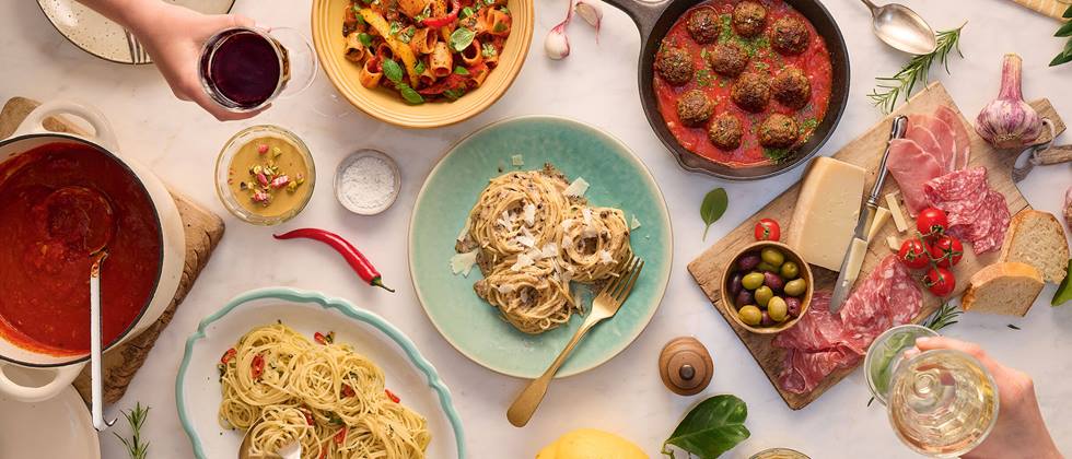 Das Bild zeigt eine von oben aufgenommene Ansicht eines reichhaltig gedeckten Tisches mit verschiedenen italienischen Gerichten. Es gibt verschiedene Pastagerichte (u.a. Spaghetti Aglio e Olio, Pasta mit Pesto und Paprika, Spaghetti mit Trüffelsoße), Fleischbällchen in Tomatensauce, eine Schüssel mit Tomatensauce, eine Aufschnittplatte mit Salami, Schinken und Käse, Oliven und Brot. Zwei Personen sind zu sehen, die gerade von den Gerichten essen. Rot- und Weißwein stehen ebenfalls auf dem Tisch. Das Ganze wirkt sehr opulent und appetitanregend, typisch für ein italienisches Festmahl.