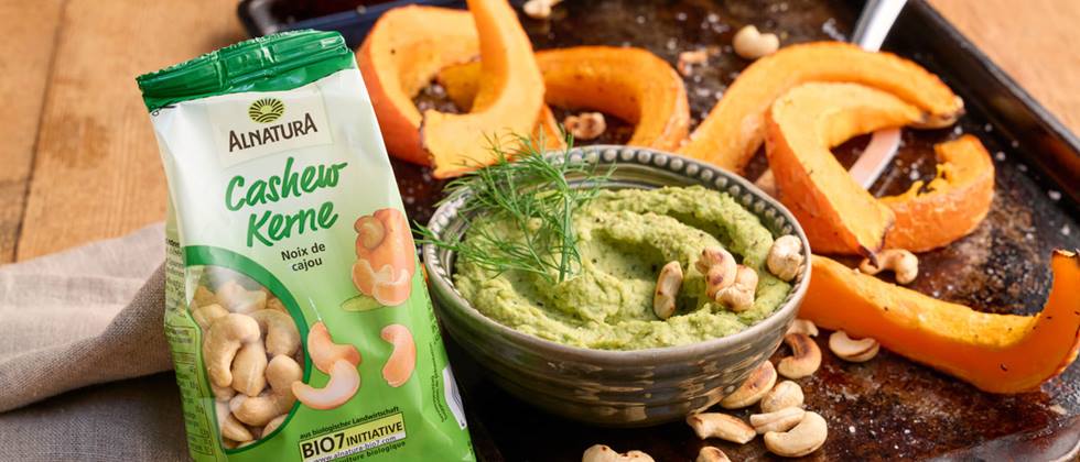 Eine Packung Alnatura Cashew-Kerne steht vor einem schwarzen Backblech mit gebackenen Hokkaido-Kürbisspalten. Auf dem Backbleck liegen verstreut geröstete Cashew-Kerne und eine kleine Porzellanschüssel mit einem grünen Mus steht darauf. Neben dem Blech ist noch eine braune Stoffserviette.