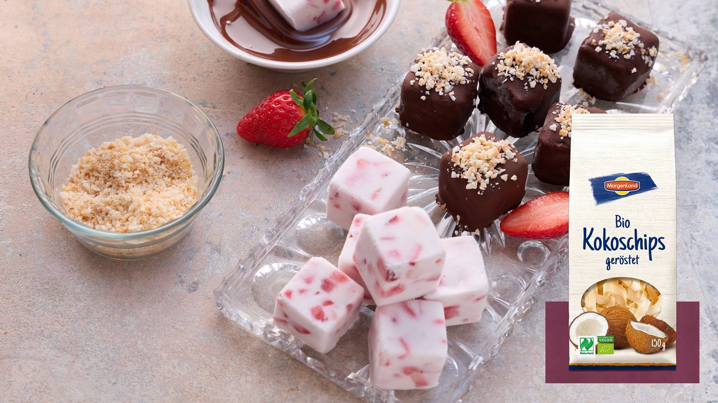 Auf einem Glasteller liegen zwei Arten selbst gemachtes Eiskonfekt mit Erdbeeren. Die eine Art ist ohne Schokoladen-Überzug die andere mit Schokoladen-Überzug und Streusel garniert. Auf dem Teller liegen zusätzlich aufgeschnittene Erdbeeren. Neben dem Glasteller steht eine Schüssel mit geraspelten Kokoschips geröstet und im Hintergrund liegt eine ganze Erdbeere und es steht eine Schüssel mit flüssiger Schokolade in der gerade eine Eiskonfekt eingetaucht wird. Ebenso ist eine Tüte Bio Kokoschips geröstet von Morgenland abgebildet.
