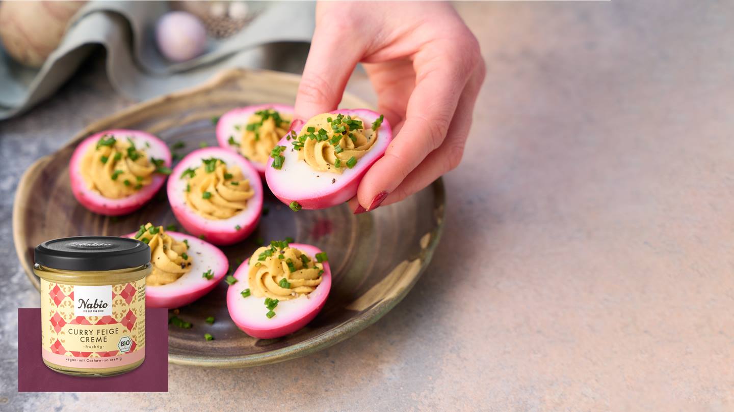 Das Rezept des Monats April sind pinke Eier mit einer Creme-Füllung. Diese liegen auf einem dunklen Teller vor einer türkisen Stoff-Serviette. Die Creme ist mit Schnittlauch garniert. Ein Ei wird von einer Hand mit roten Fingernägeln in die Kamera gehalten. Zudem ist das Produkt im Glas "Curry Feige Creme" der Marke Nabio zu sehen. Das Produkt ist fruchtig, vegan, mit Chashew und wird als so cremig angegeben. Das Etikett enthält ebenso das deutsche Bio-Siegel.