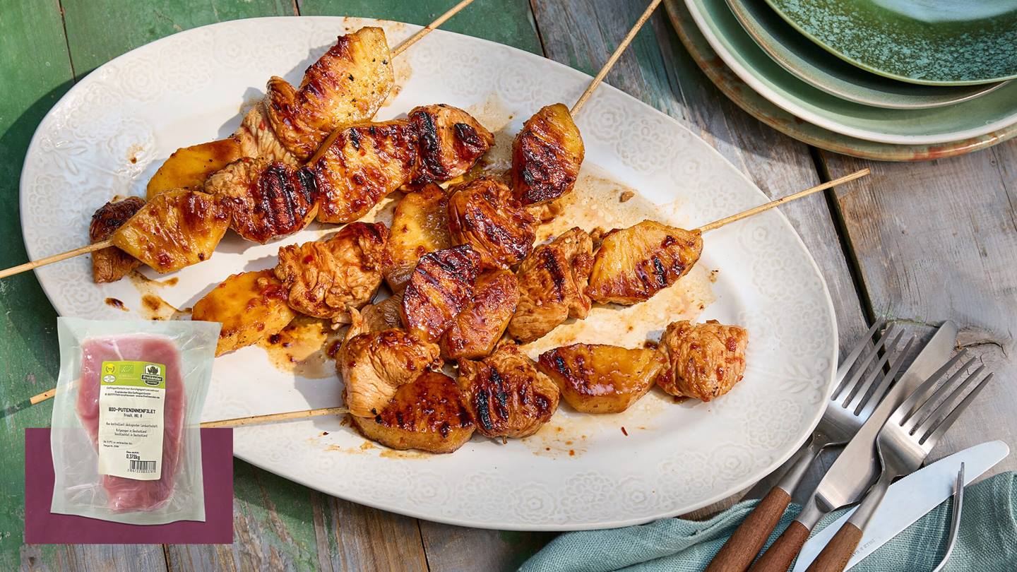 Auf einem Vintage-Holztisch steht eine weißer Teller auf diesem liegen fertige gegrillte Ananas-Puten-Spieße. Neben dem Teller liegt Besteck und eine Stoff-Serviette. Zusätzlich ist das Produkt von Freiländer Bio-Puteninnenfilet, frisch abgebildet.
