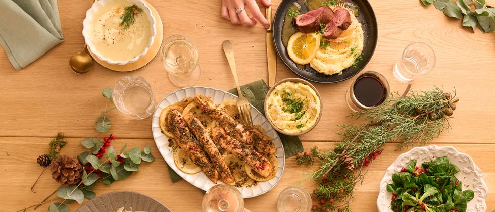Ein festlich gedeckter Holztisch mit einer Vielfalt an kulinarischen Köstlichkeiten: Links oben eine Schüssel mit cremiger Suppe, daneben ein Teller mit zartem Fleischgericht und Zitronenscheiben, in der Mitte ein Teller mit knusprig gebackenen Fischfilets und Zitronenscheiben, unten links ein Teller mit herzhaftem Risotto, unten rechts ein frischer Salat mit grünen Blättern und Granatapfelkernen. Dazu Gläser mit Wein und Wasser, goldfarbenes Besteck und dekorative Elemente wie Tannenzapfen, Eukalyptuszweige und Beeren.